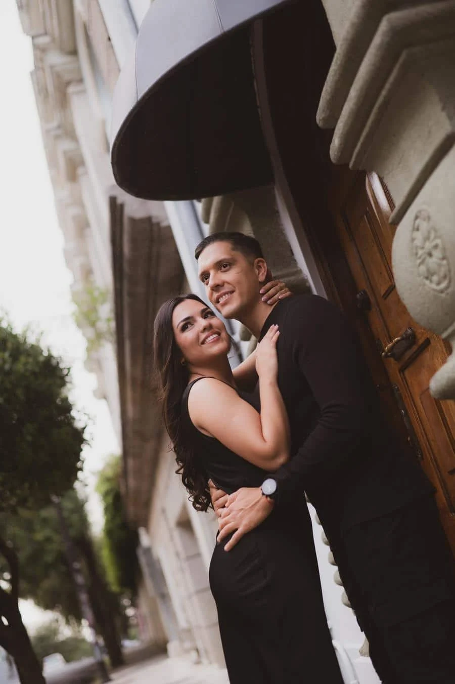 fotografía-de-compromiso-costa-rica-15 | Pacific Photography Pareja con atuendos negros abrazándose y sonriendo afuera de una puerta ornamentada en una calle de la ciudad.