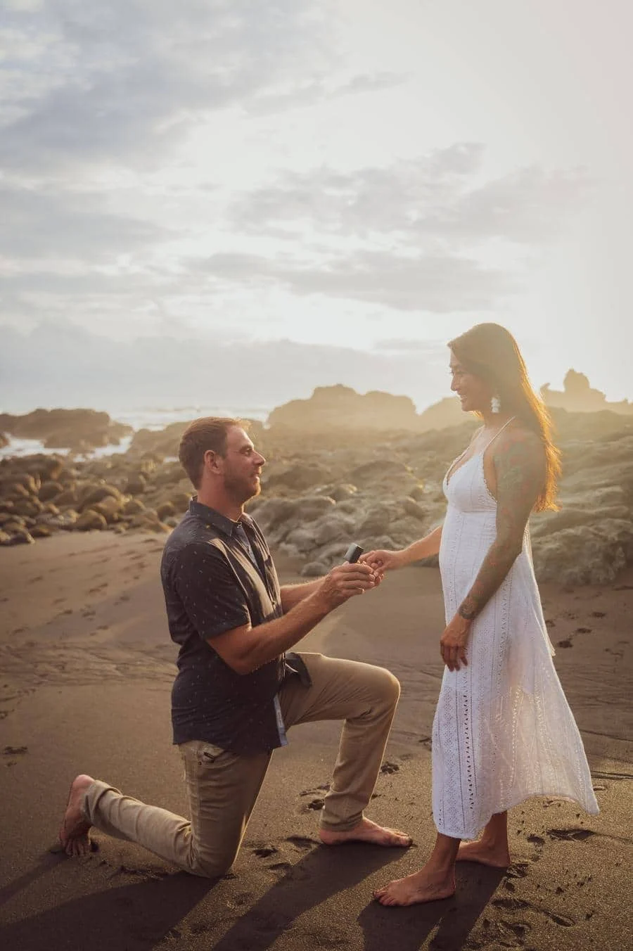 engagement photography costa rica-3 | Pacific Photography Man kneels on a sandy beach at sunset, presenting a ring to a standing woman in a white dress.