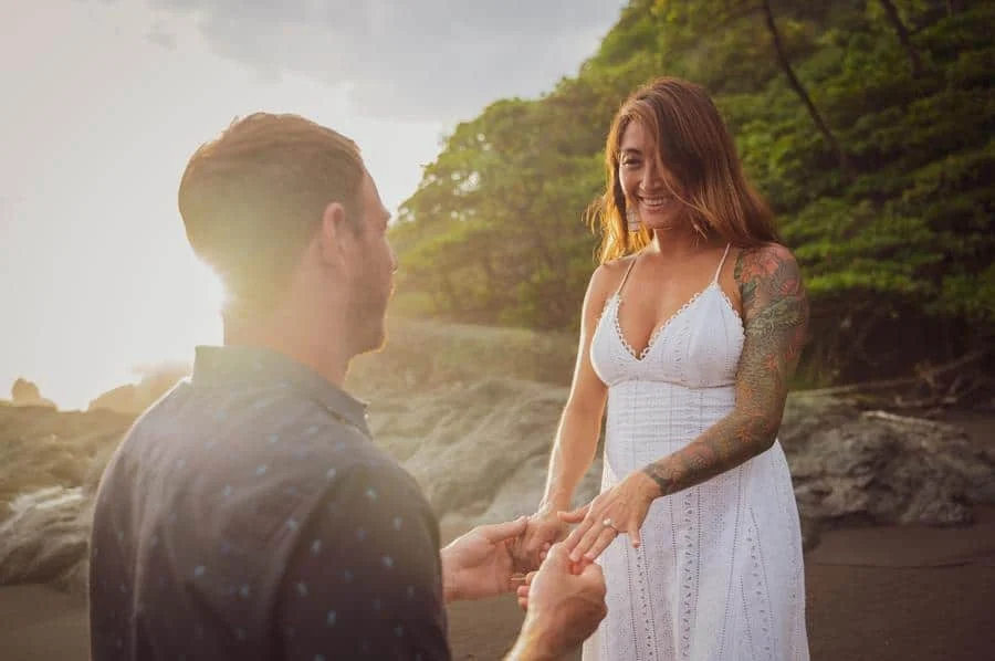 engagement photography costa rica-4 | Pacific Photography Man proposing on a sunlit beach, placing a ring on a woman's finger as she smiles.