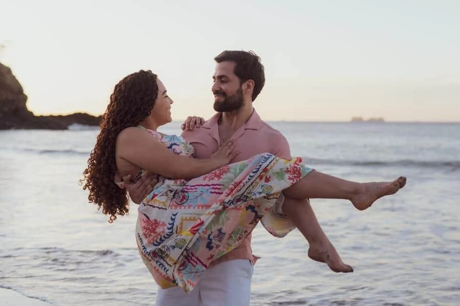 fotografía-de-compromiso-costa-rica-8 | Pacific Photography Pareja en una playa al atardecer; el hombre carga a la mujer en sus brazos envuelta en una toalla/manta colorida mientras se sonríen mutuamente.