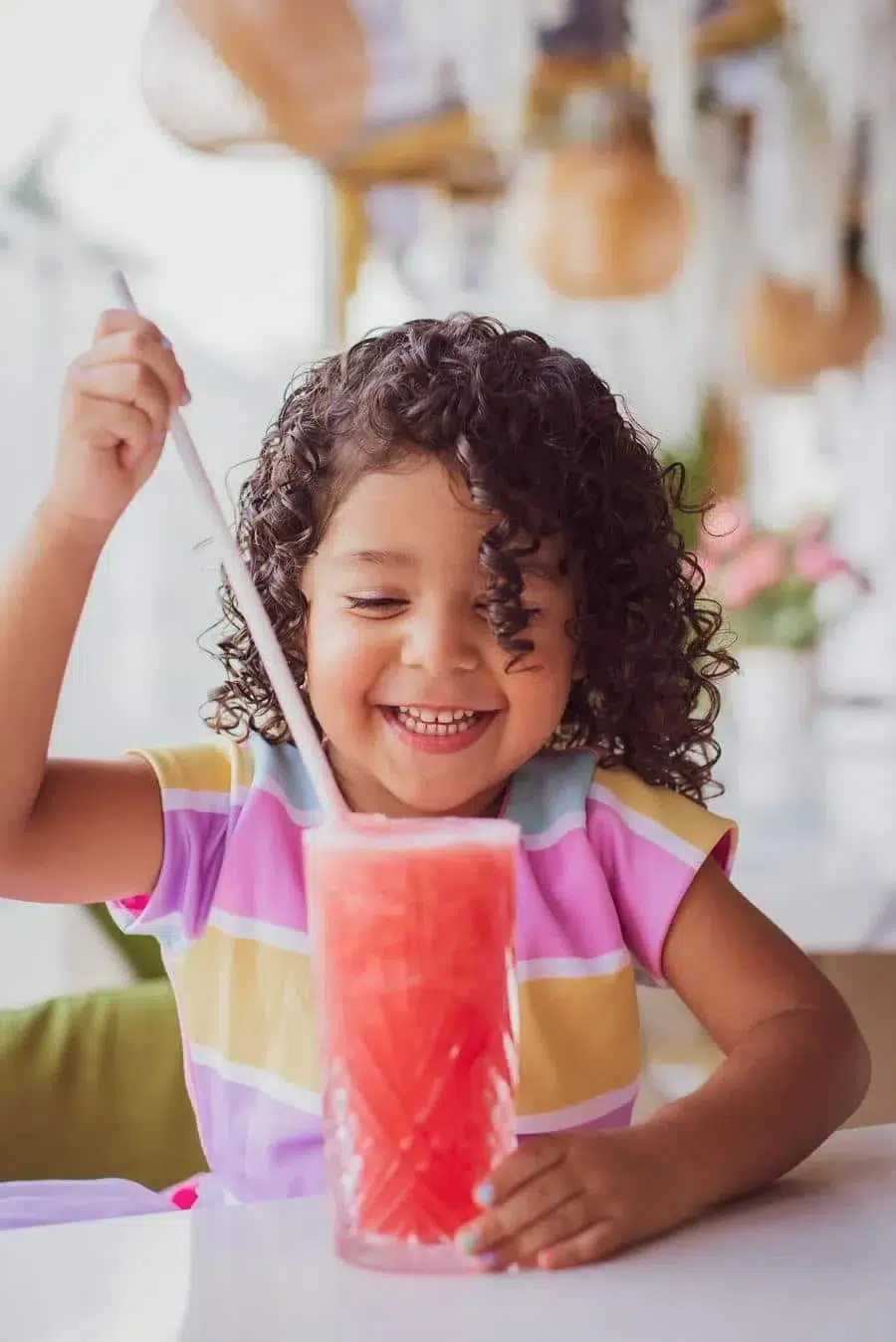 Happy young girl with curly hair enjoying a pink-red slushie with a straw at a bright table.