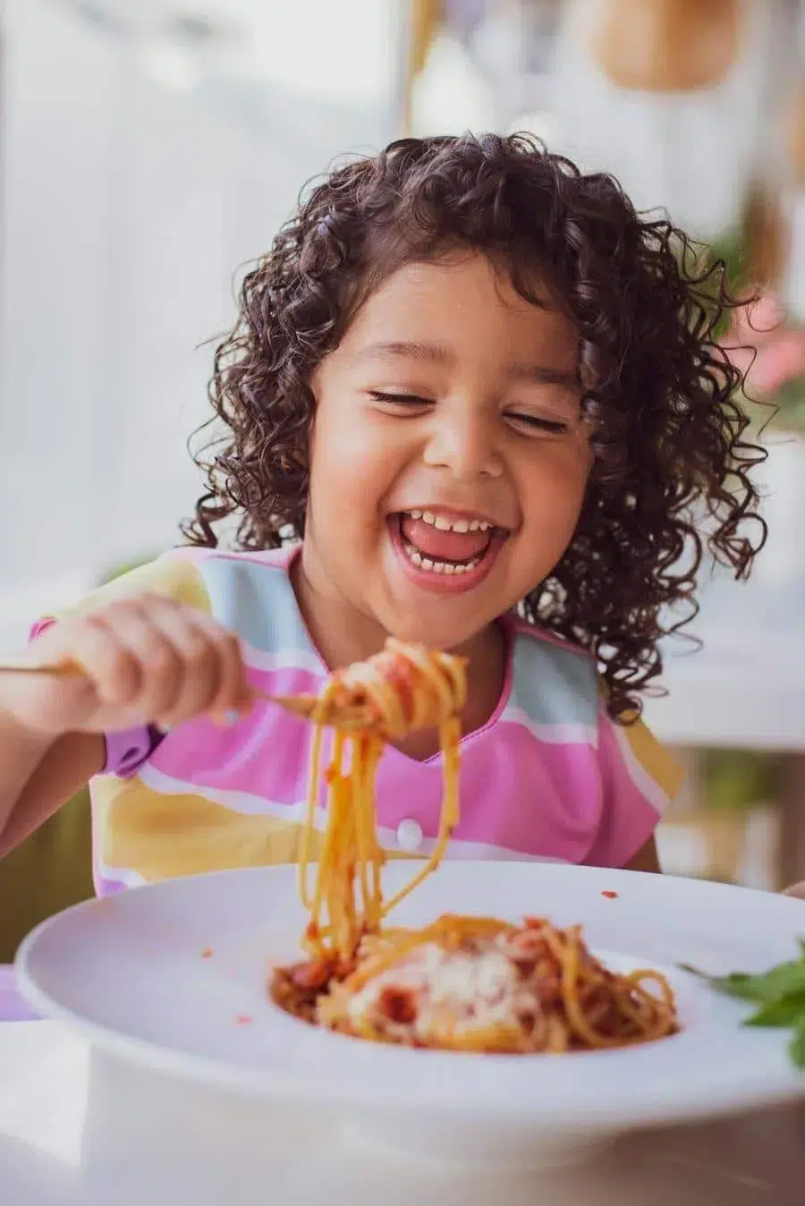 A young girl with curly hair enjoying spaghetti, twirling noodles with a big smile at the table.