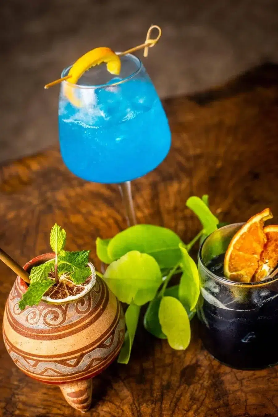 Blue tropical cocktail with orange twist in a stem glass, alongside a minty drink in a decorative clay pot and a dark cocktail with an orange garnish on a wooden table in the background.