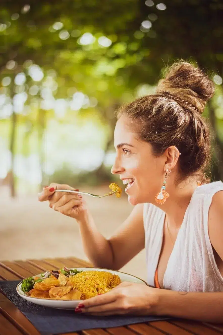 Woman eating outdoors at a wooden table, enjoying a plate of yellow rice with vegetables in a park setting