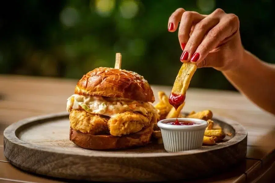 food menu and restaurant photography costa rica -16 | Pacific Photography Crispy chicken sandwich on a glossy bun with fries and a small cup of ketchup on a wooden board; a hand with red nails dips a fry into the ketchup.