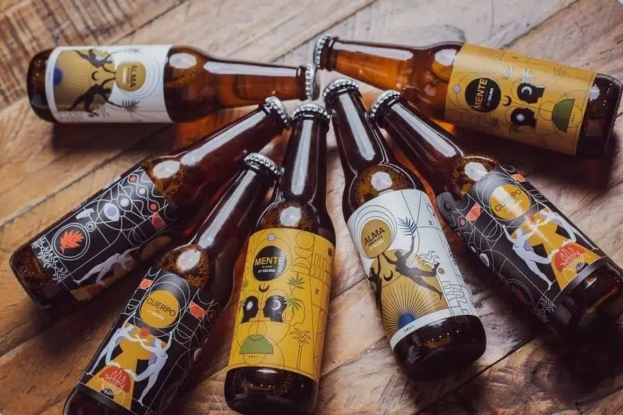 food menu and restaurant photography costa rica -18 | Pacific Photography Several brown beer bottles arranged in a circular cluster on a wooden surface, each with colorful illustrated labels featuring tropical or geometric designs and text like 'MENTE', 'ALMA', and 'CUID?