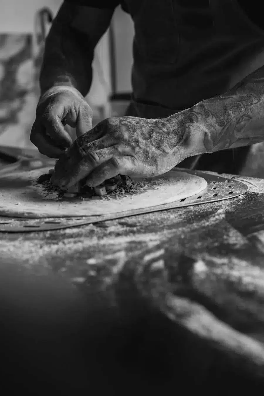 Tattooed hands shaping dough on a flour-dusted pizza surface.