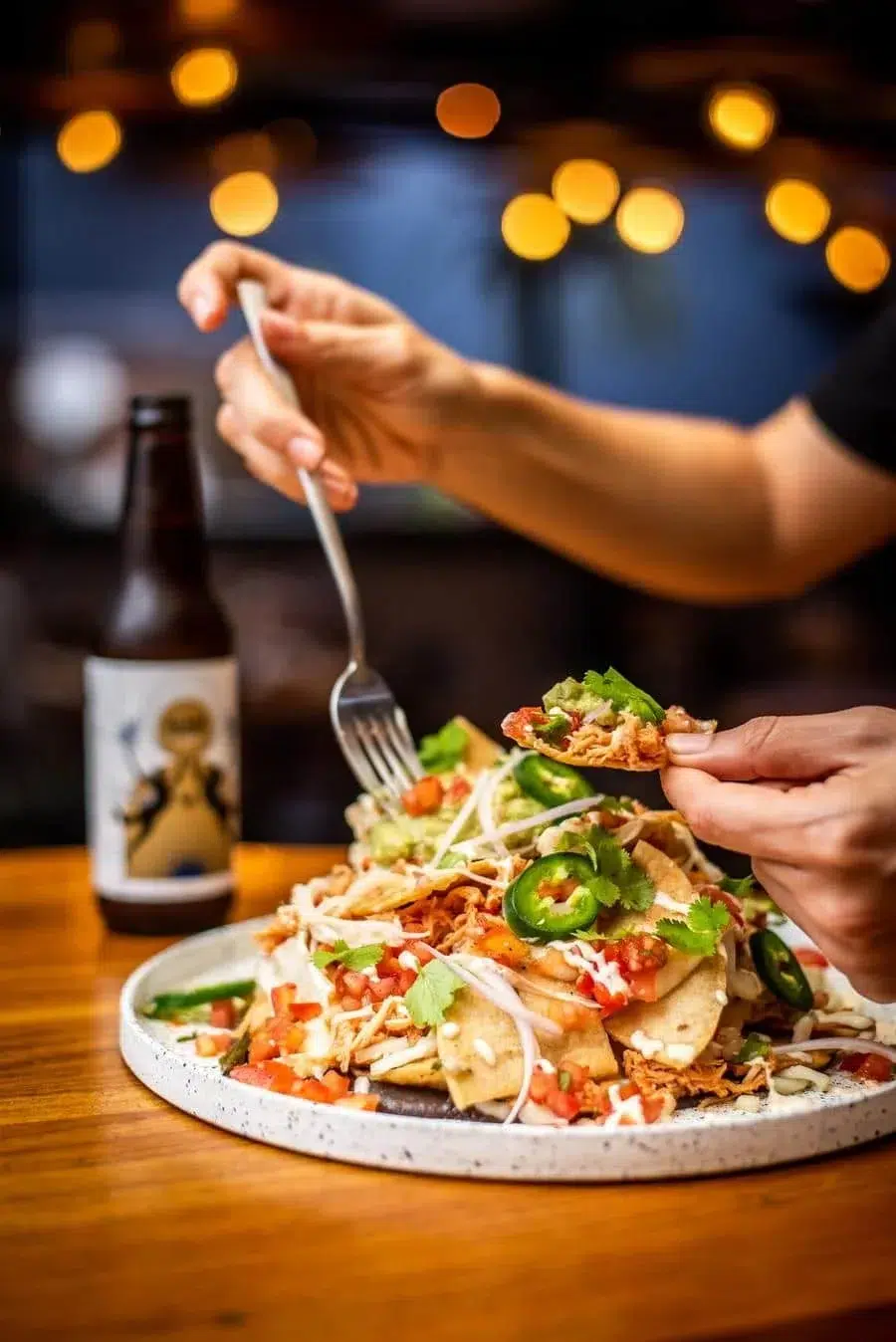 Person assembling a colorful taco plate with cilantro, jalapeños, tomatoes and slices of tortilla on a ceramic platter; a beer bottle sits in the blurred background.