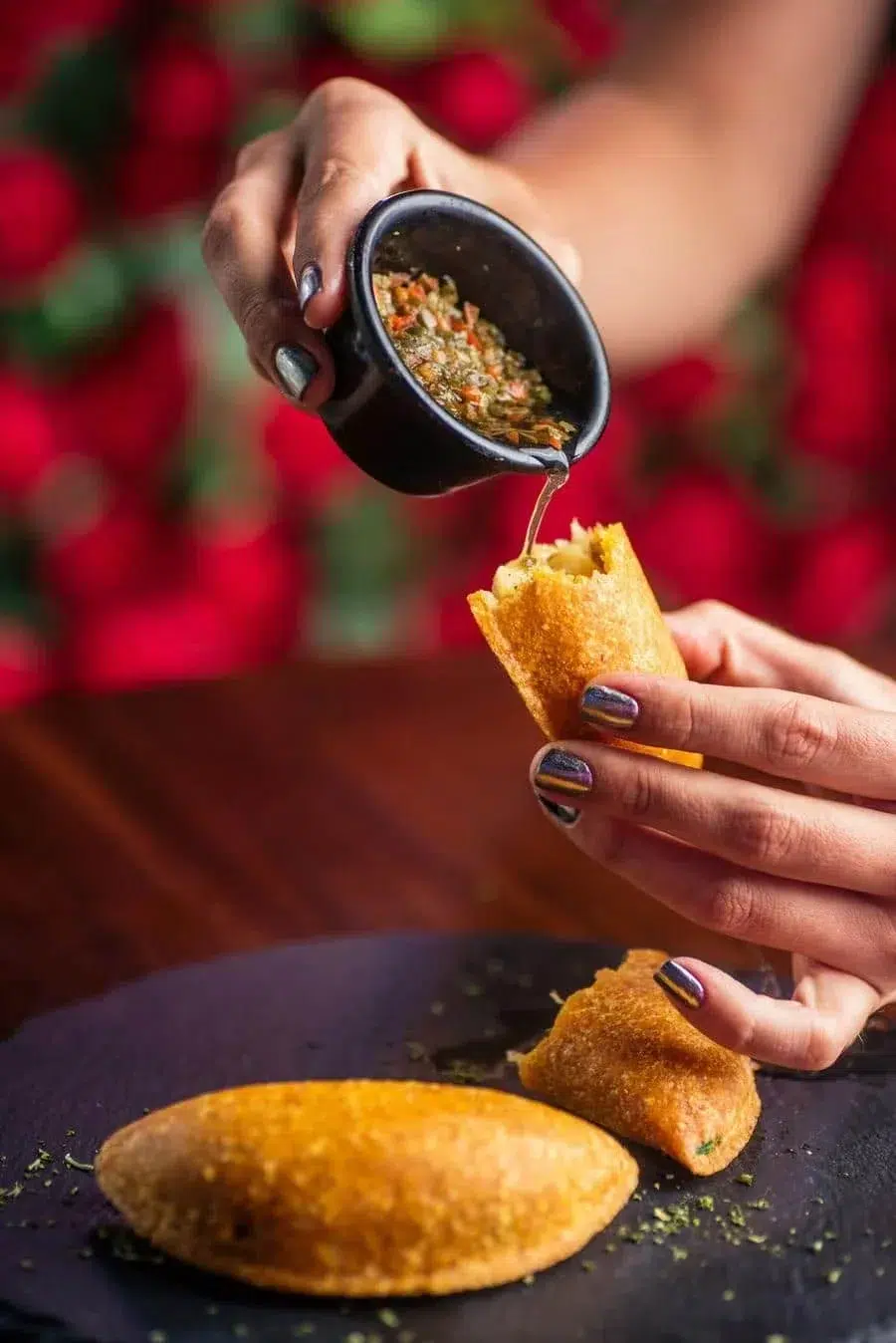 Hands pouring a green herb sauce from a small black pitcher onto a crispy stuffed pastry held in the other hand.