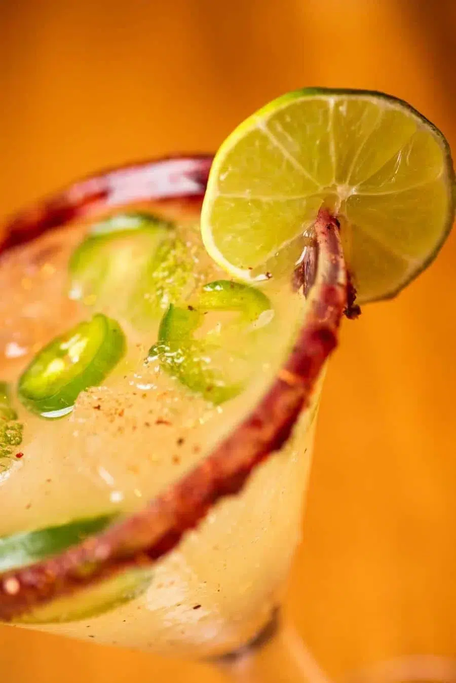 Margarita glass with a lime wedge, jalapeño slices, and a salted rim against an orange background.