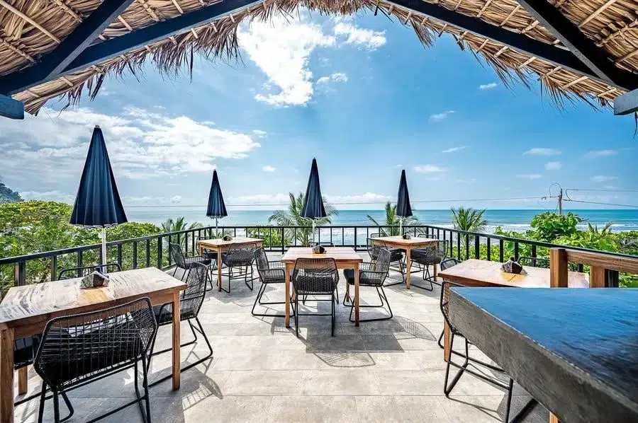food menu and restaurant photography costa rica -28 | Pacific Photography Outdoor seaside cafe terrace with wooden tables, black chairs, blue umbrellas, and an ocean-view backdrop.
