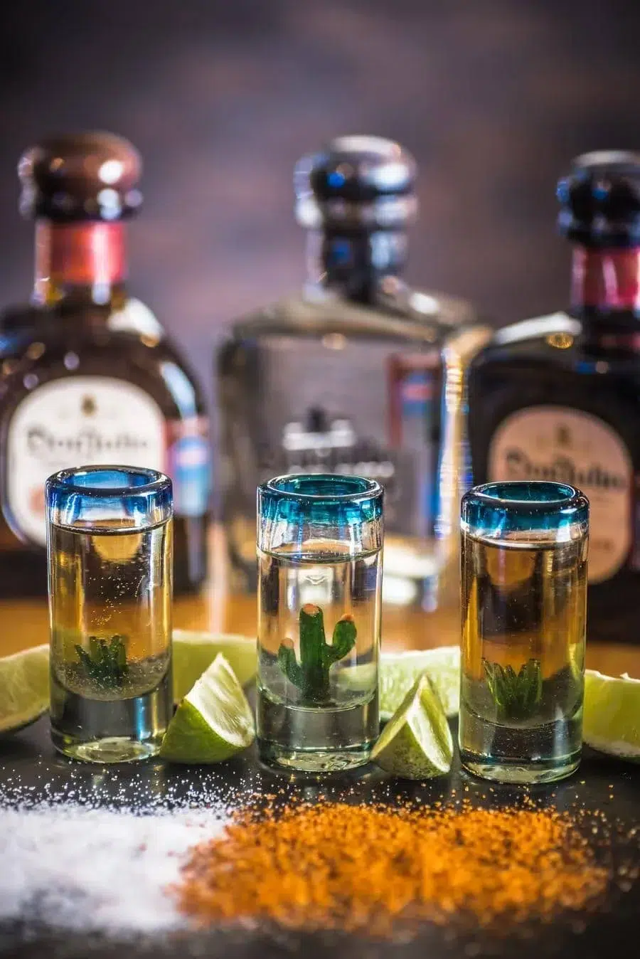 Three blue-rimmed shot glasses with cactus garnishes and lime wedges on a bar, tequila bottles blurred behind them.