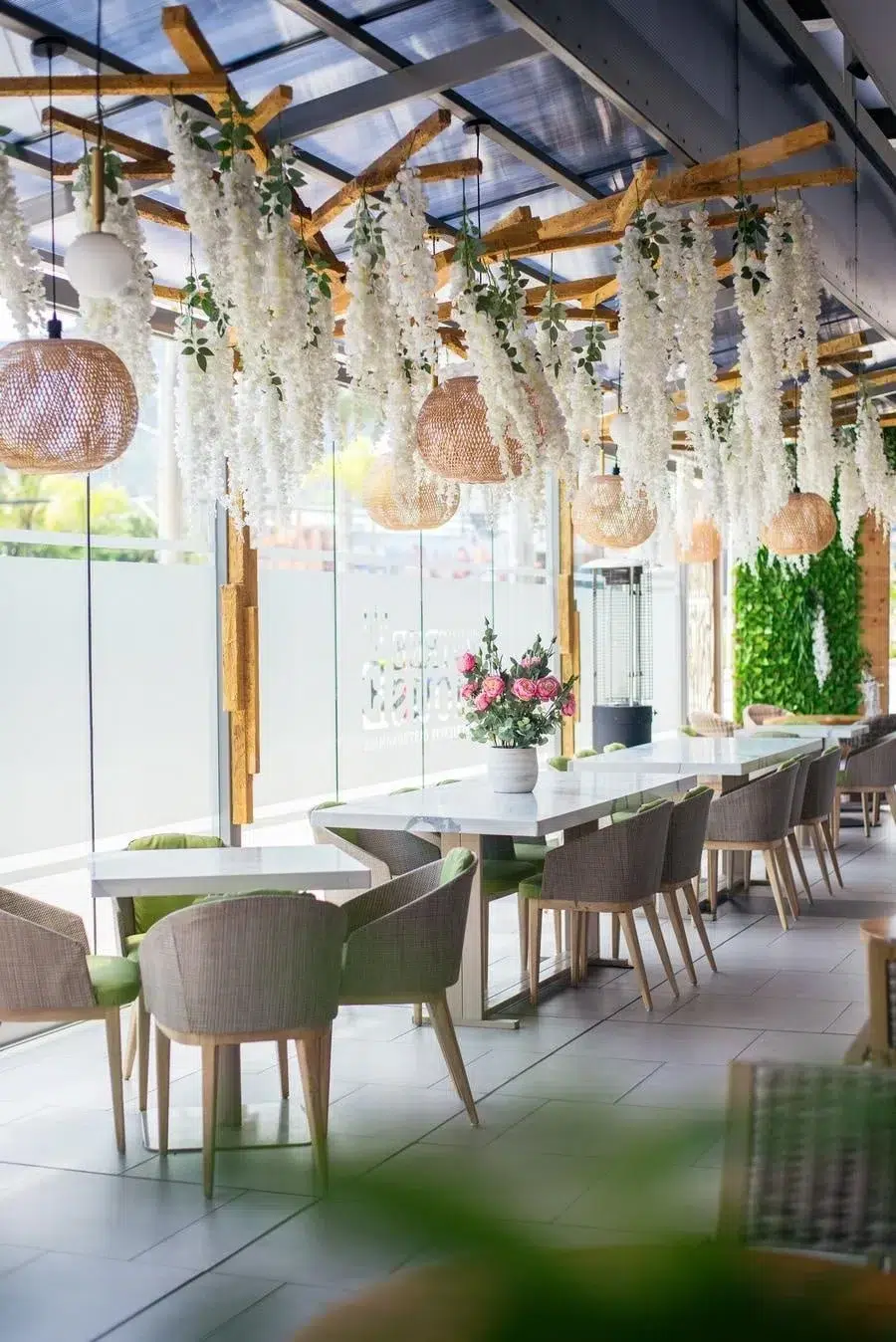 Bright modern cafe interior with white hanging flowers, wicker pendant lights, glass walls, and green chairs.