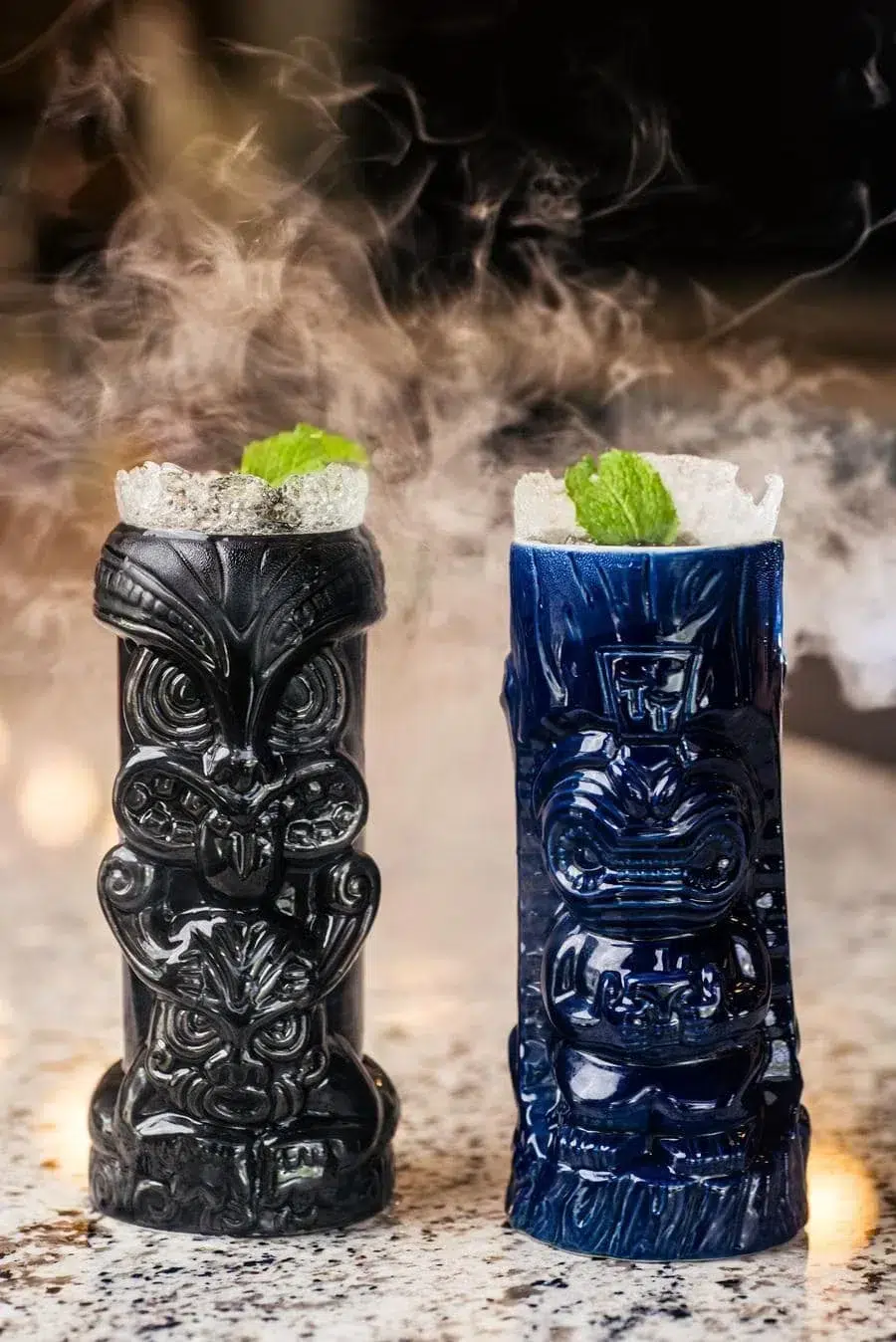 Two carved tiki mugs (black on left, blue on right) filled with crushed ice and mint, with wisps of steam rising between them on a speckled counter.