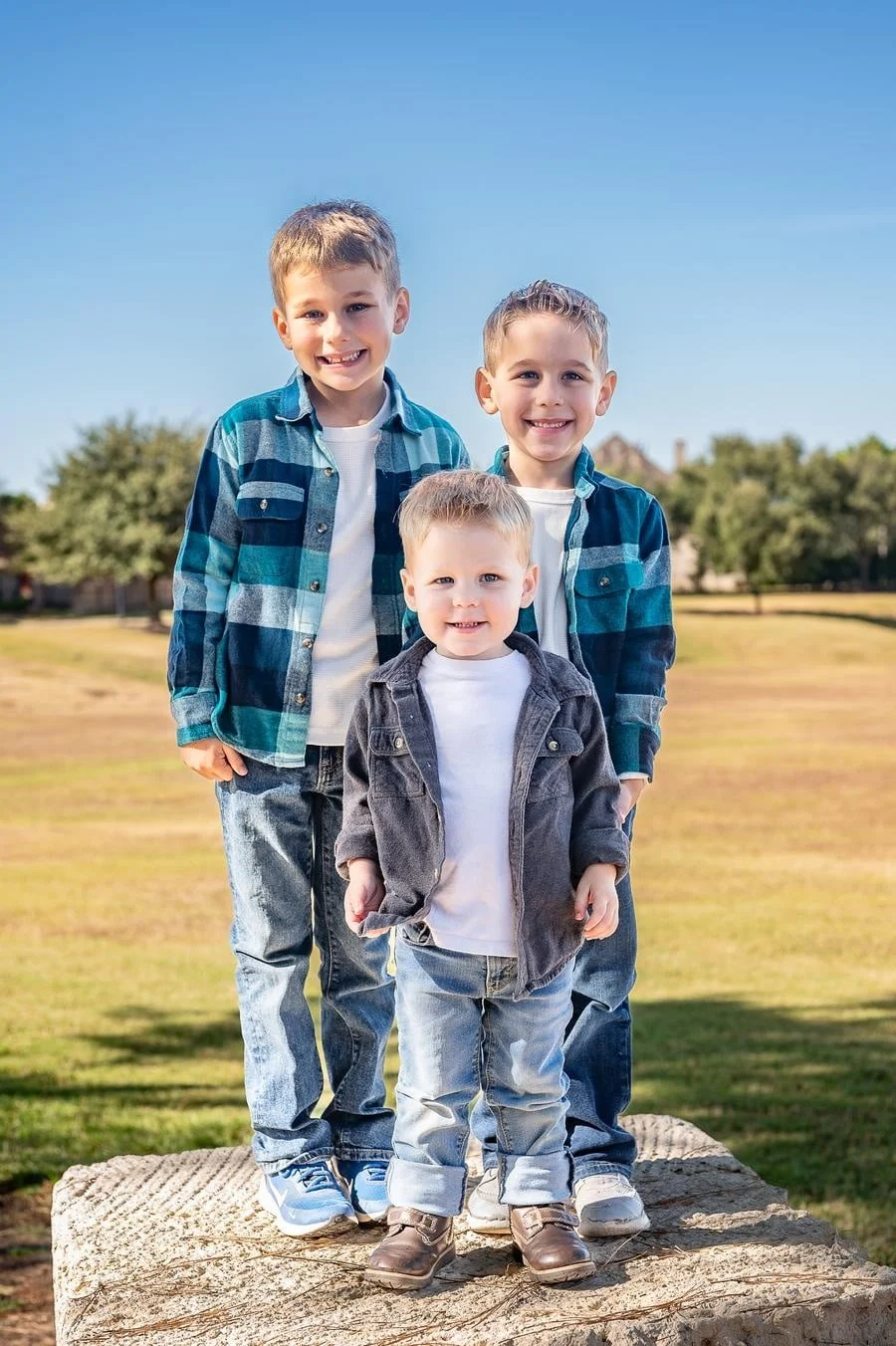 people and lifestyle photography -1 | Pacific Photography Three smiling boys pose together on a large rock outdoors on a sunny day, wearing denim jackets and white shirts with a blue sky behind them.
