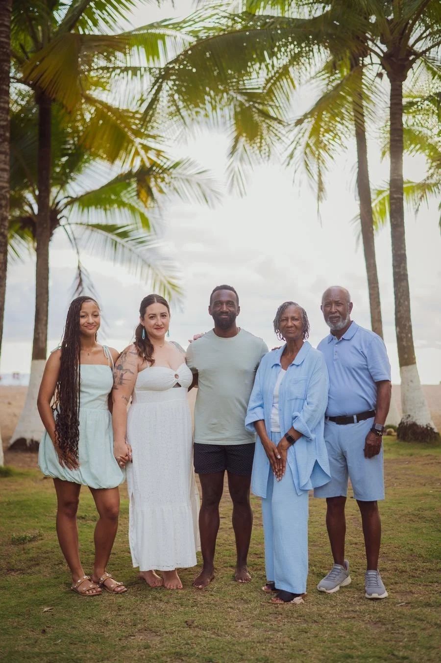 people and lifestyle photography -10 | Pacific Photography A family of five stands together on a grassy beach under palm trees, posing for a group photo in light summer outfits.