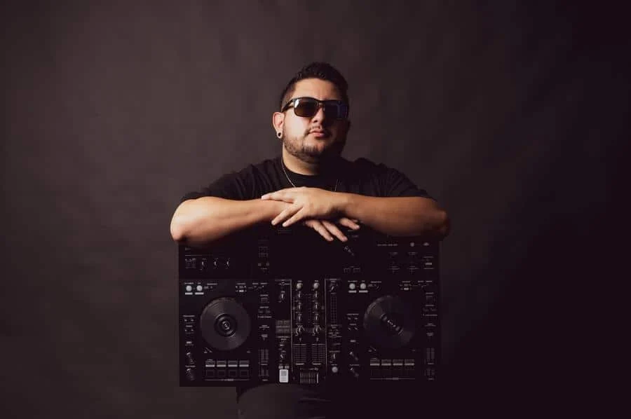 people and lifestyle photography -12 | Pacific Photography DJ wearing sunglasses rests his arms on a large professional mixer against a dark backdrop.