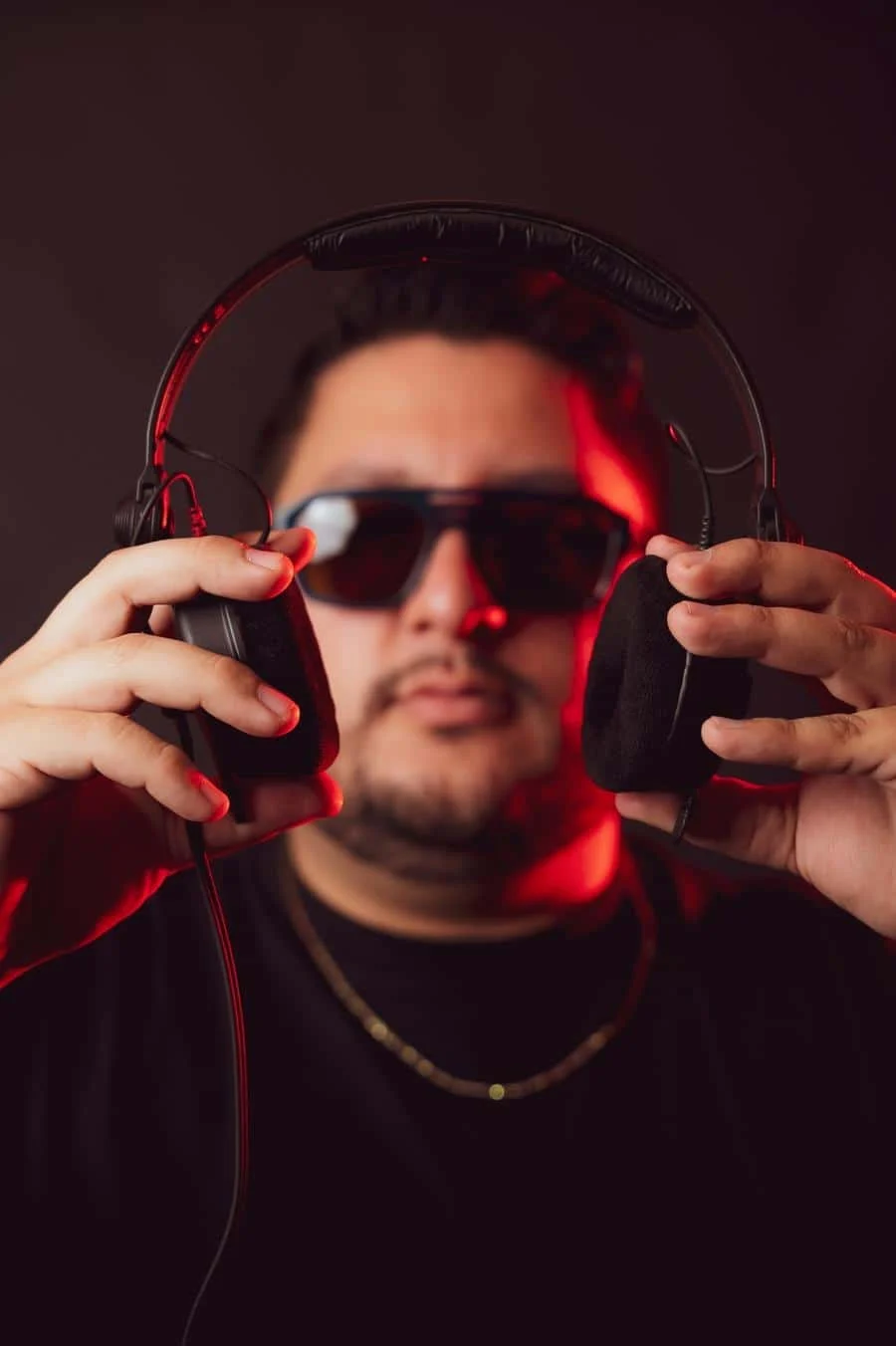 people and lifestyle photography -16 | Pacific Photography DJ wearing sunglasses and headphones, holding the ear cushions toward the camera with red lighting in the background