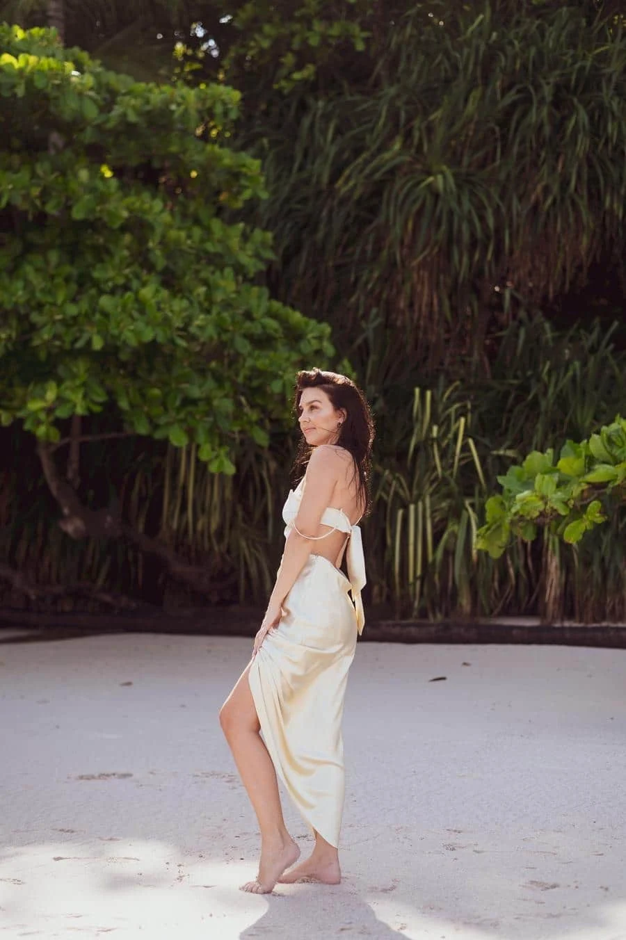 people and lifestyle photography -17 | Pacific Photography Woman in a white satin dress standing on a sandy beach, tropical greenery behind her, looking over her shoulder.