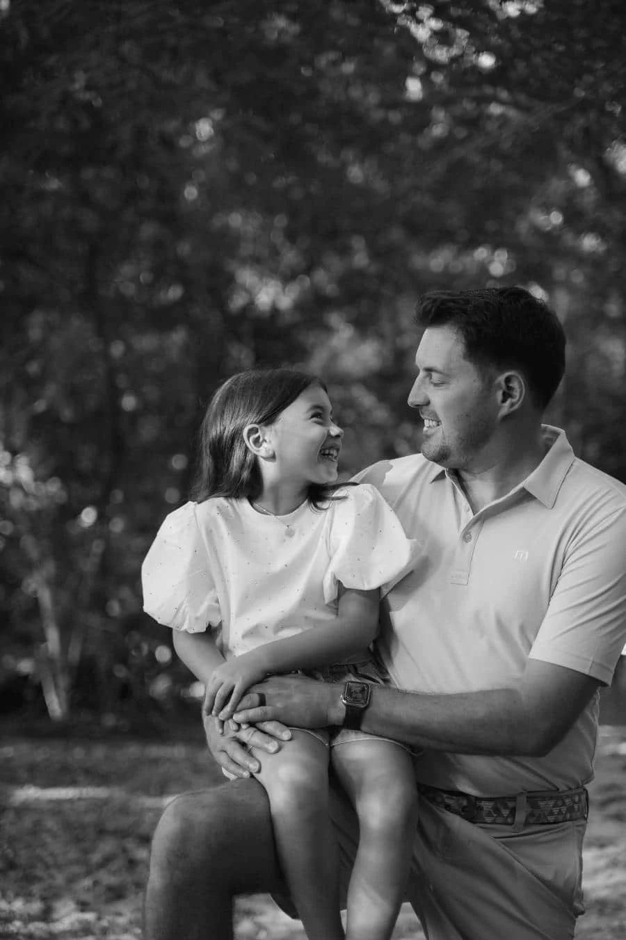 people and lifestyle photography -18 | Pacific Photography Father sitting outdoors with his young daughter on his lap, smiling at each other in a park (black and white).