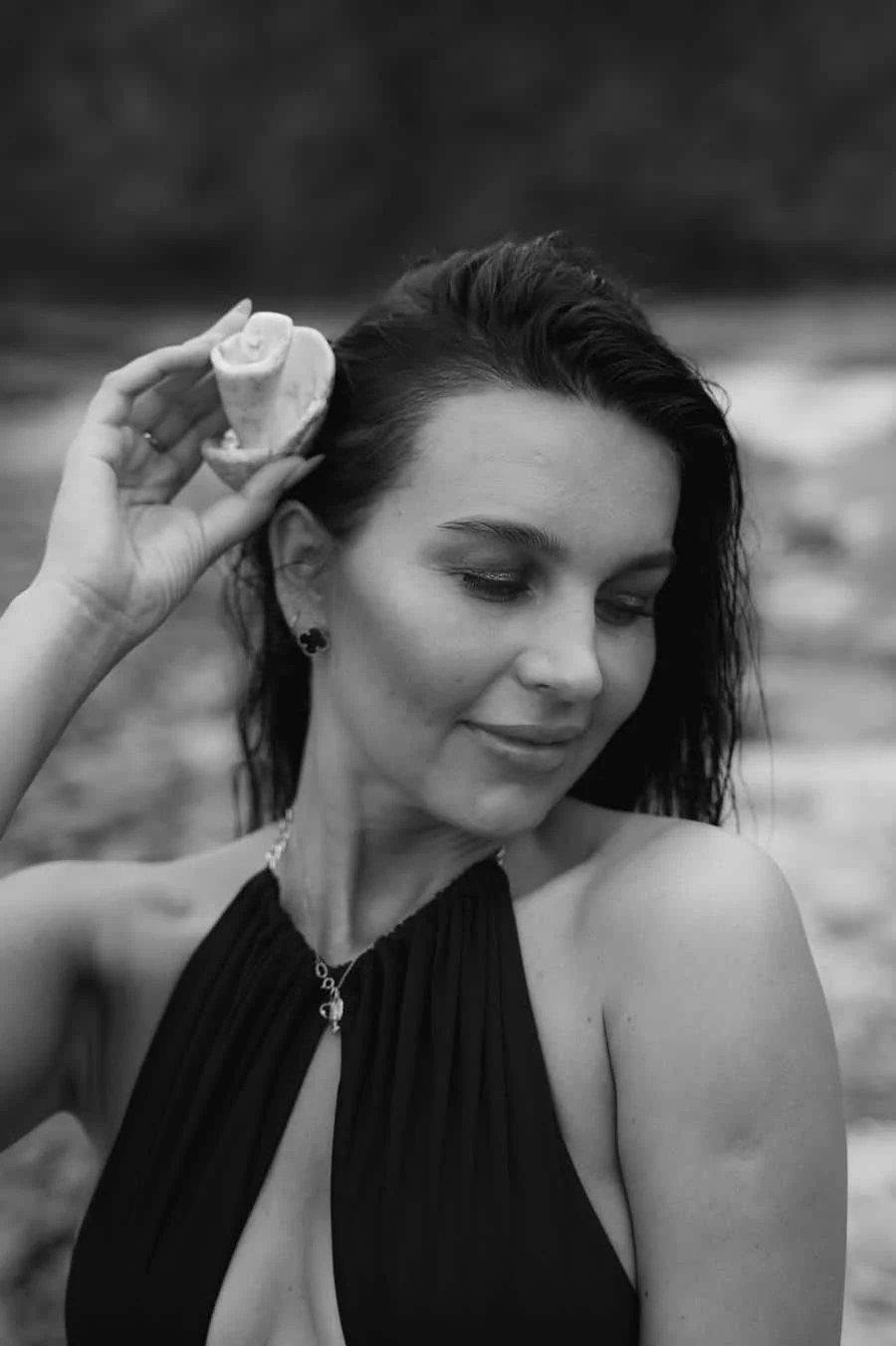 people and lifestyle photography -19 | Pacific Photography Woman in a black halter top holds a seashell to her ear as if listening, outdoors in grayscale.