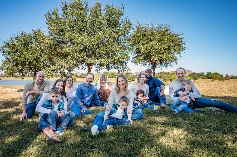 people and lifestyle photography -2 | Pacific Photography A large family sits and smiles on the grass beneath trees, with a lake in the background on a sunny day.