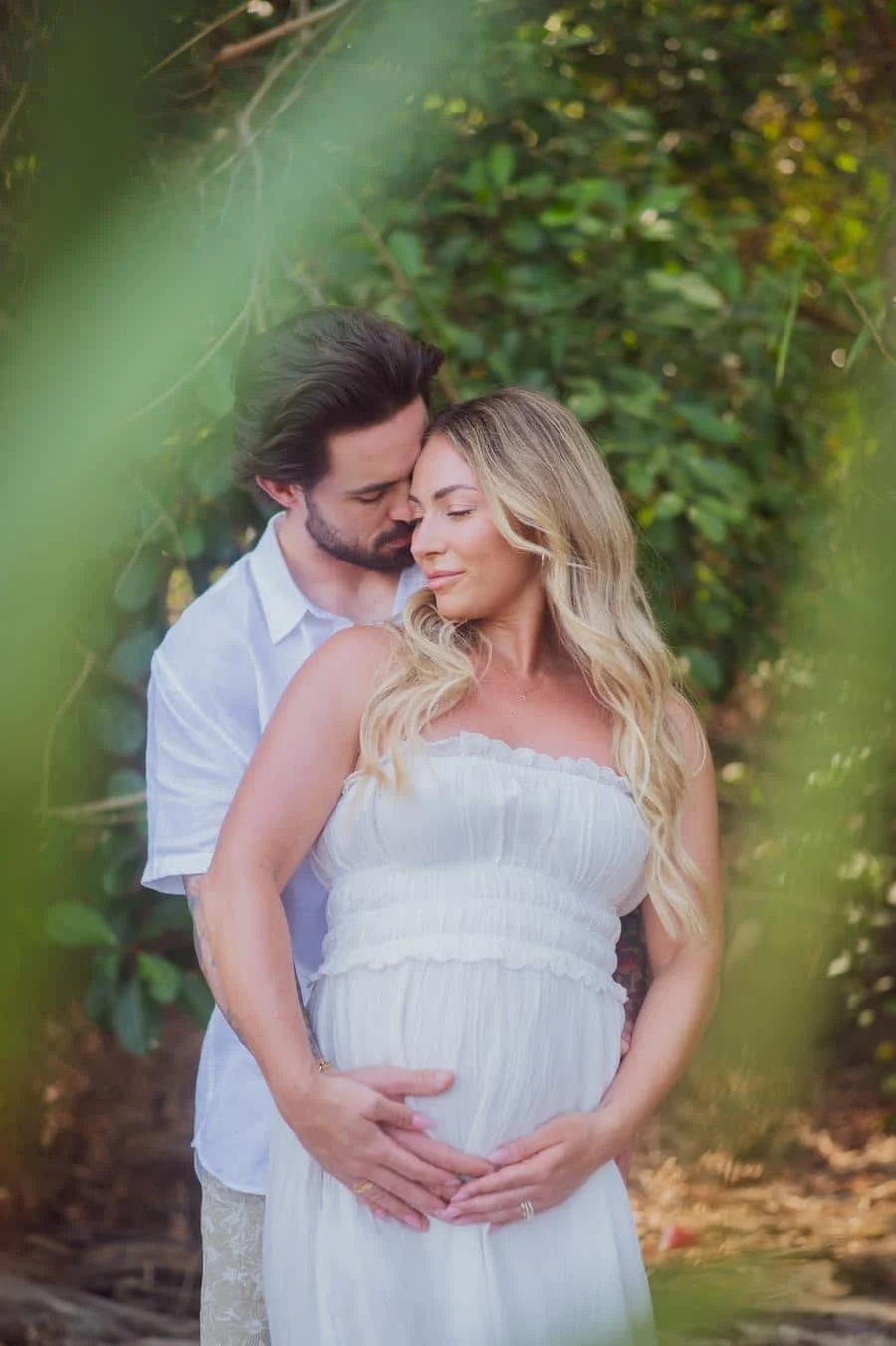 people and lifestyle photography -21 | Pacific Photography Pregnant woman in a white dress stands outdoors as her partner hugs her from behind, hands on her baby bump in a green garden setting.