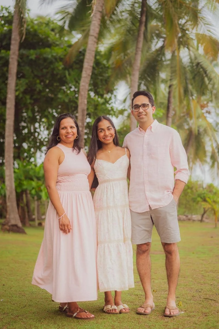 people and lifestyle photography -22 | Pacific Photography Three people posing together outdoors on a grassy area with palm trees, smiling at the camera in light pink and white outfits.