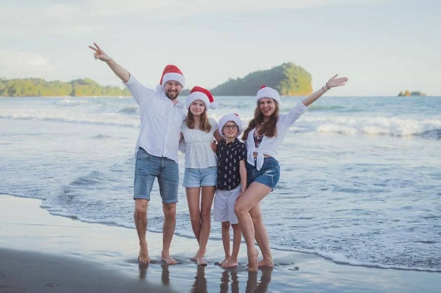 people and lifestyle photography -24 | Pacific Photography Family of four in Santa hats posing on a beach with waves in the background, smiling and arms around each other.