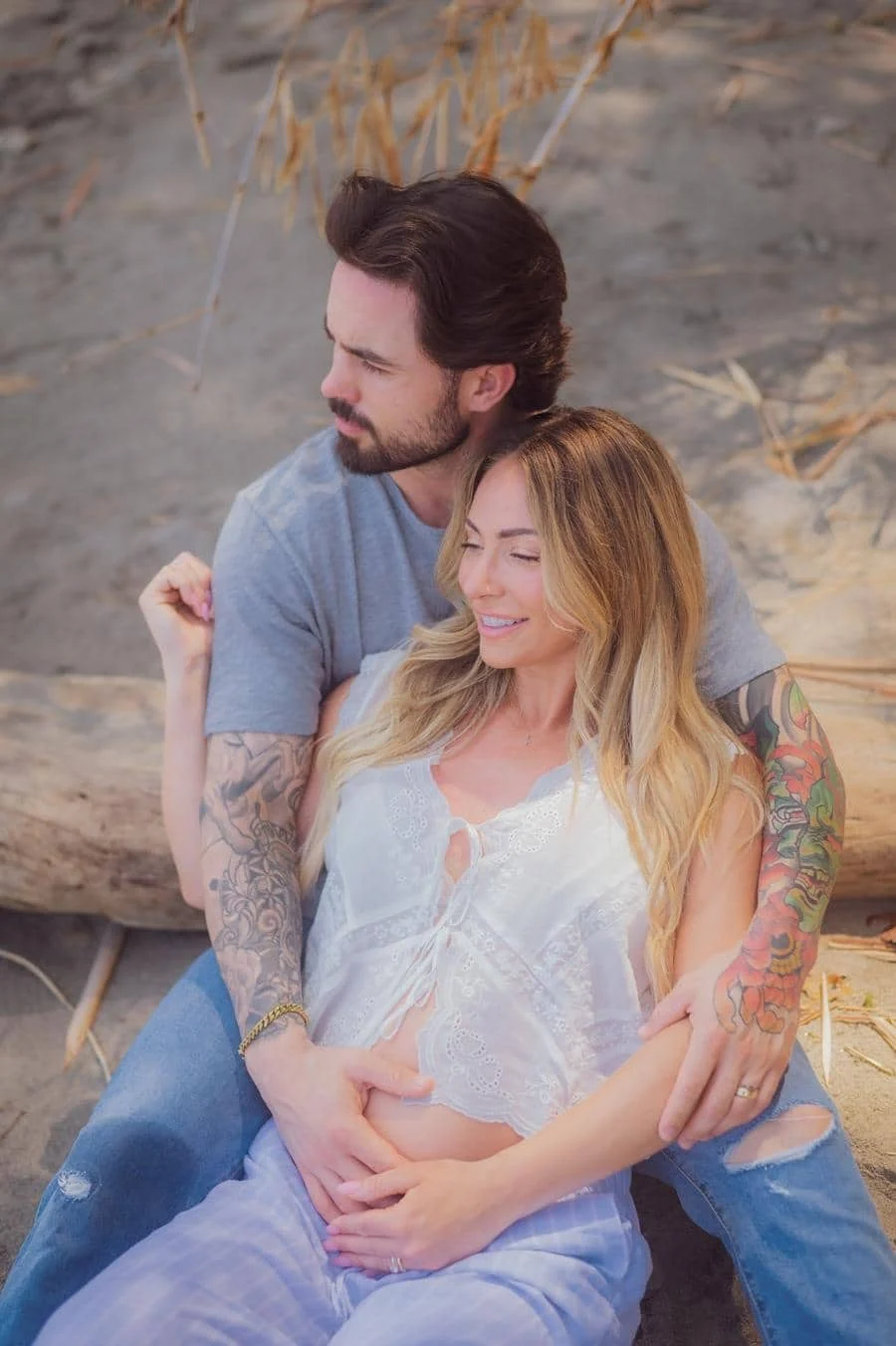 people and lifestyle photography -25 | Pacific Photography Pregnant woman in a white lace top sits on a sandy beach, embraced by a tattooed man as they smile softly.