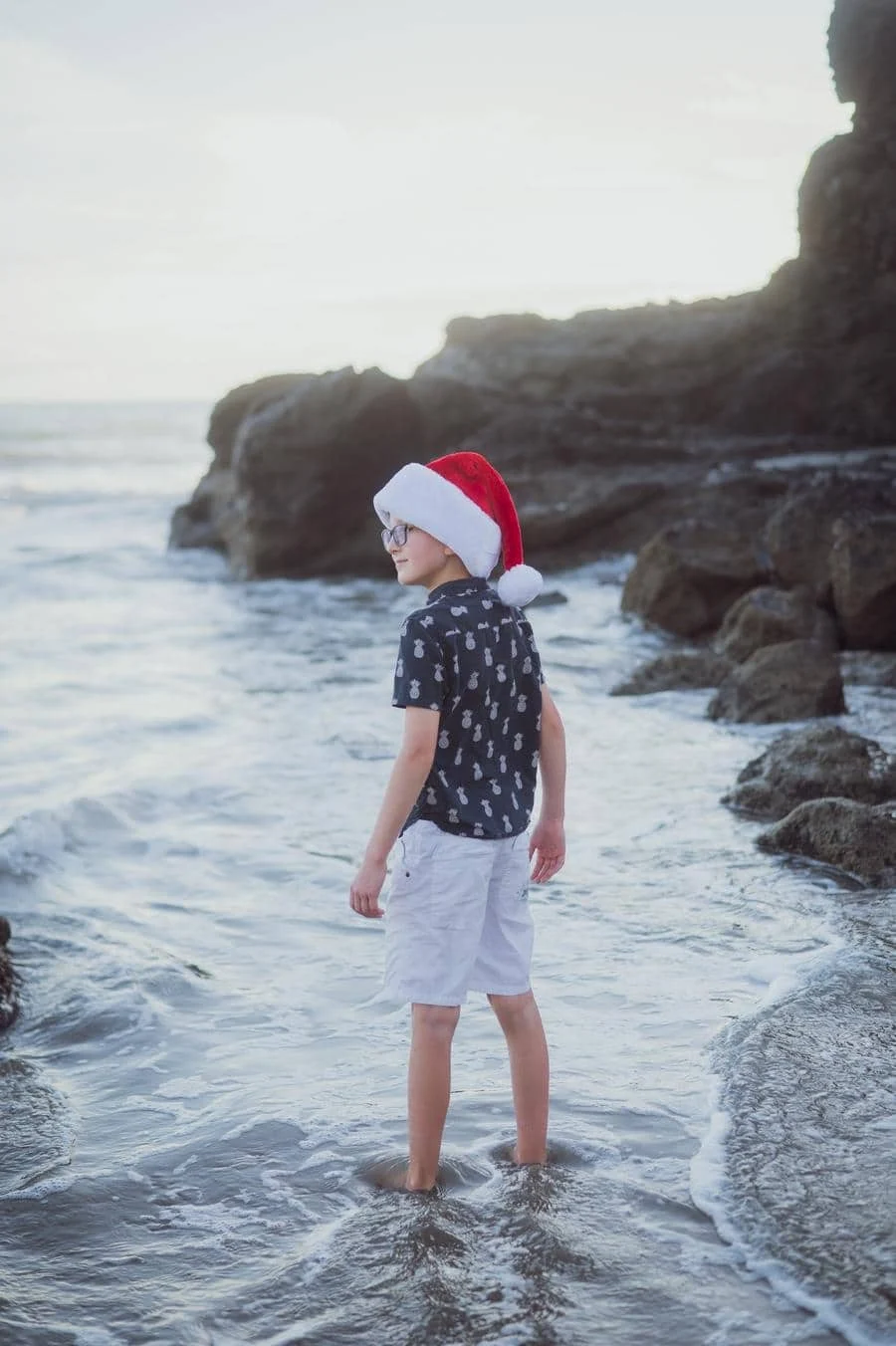 people and lifestyle photography -26 | Pacific Photography Child in a Santa hat stands barefoot in shallow water along a rocky beach.