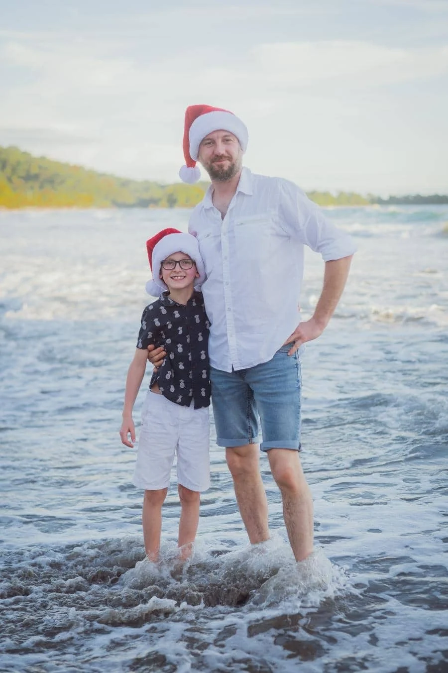 people and lifestyle photography -27 | Pacific Photography Father and son in Santa hats posing together at the beach with waves at their feet, smiling.