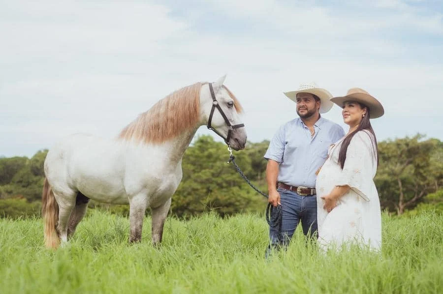 people and lifestyle photography -29 | Pacific Photography Pregnant woman and partner in cowboy hats standing with a white horse in a green field.