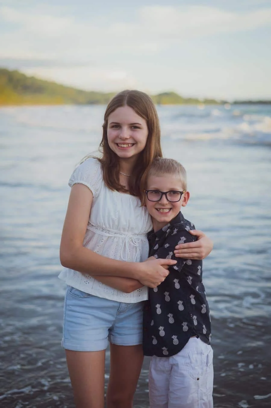 people and lifestyle photography -30 | Pacific Photography Two smiling siblings hug at the beach with calm water in the background.