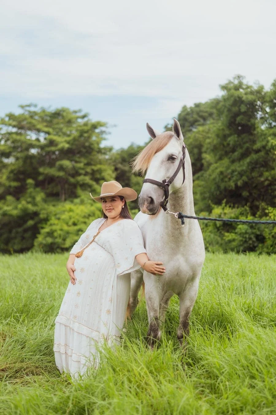 people and lifestyle photography -31 | Pacific Photography Pregnant woman in a white flowing dress and cowboy hat stands in a green field, resting a hand on her belly beside a white horse.