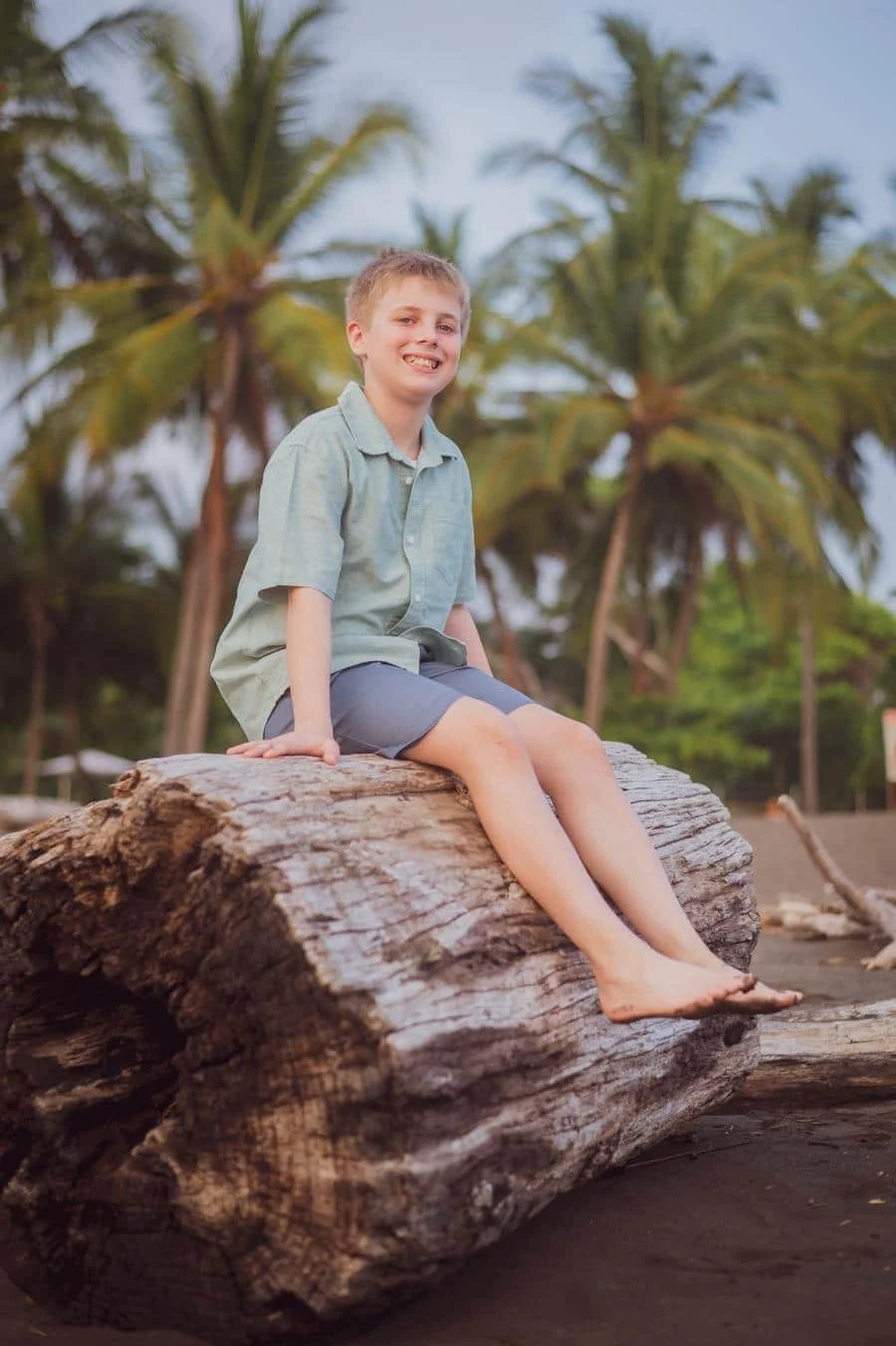 people and lifestyle photography -32 | Pacific Photography Boy sitting barefoot on a large driftwood log at a palm‑fringed beach, smiling at the camera.