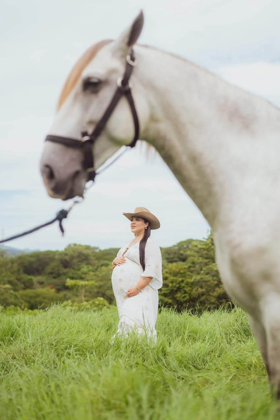 people and lifestyle photography -33 | Pacific Photography Pregnant woman in a white dress and wide-brim hat standing in a green field with a large gray horse close to the camera in the foreground.