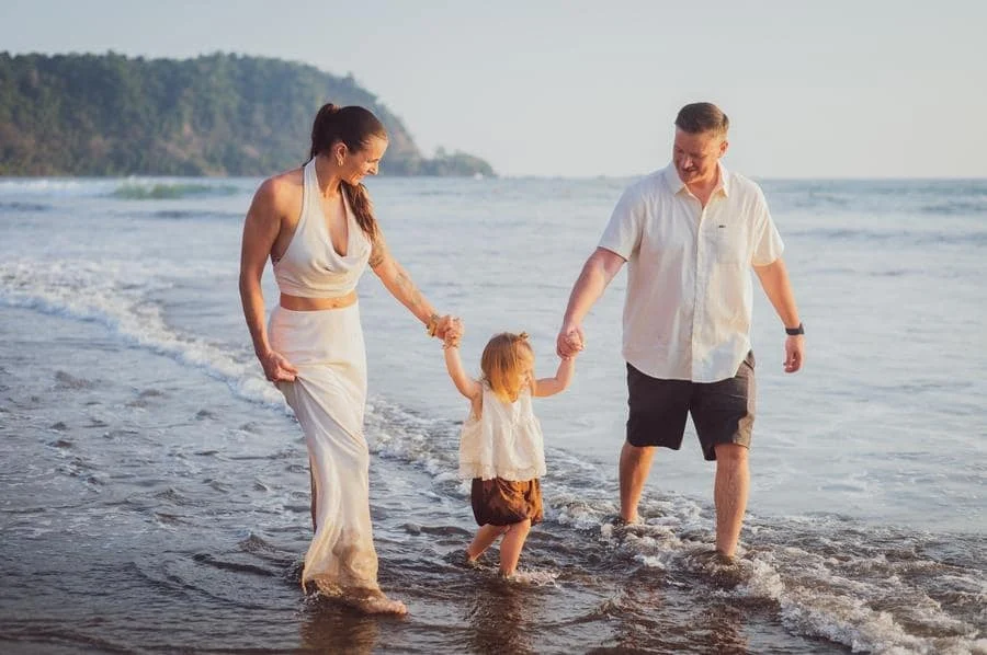 people and lifestyle photography -35 | Pacific Photography Family of three walking hand in hand along the beach shoreline with gentle waves. (Mom, dad, and toddler)