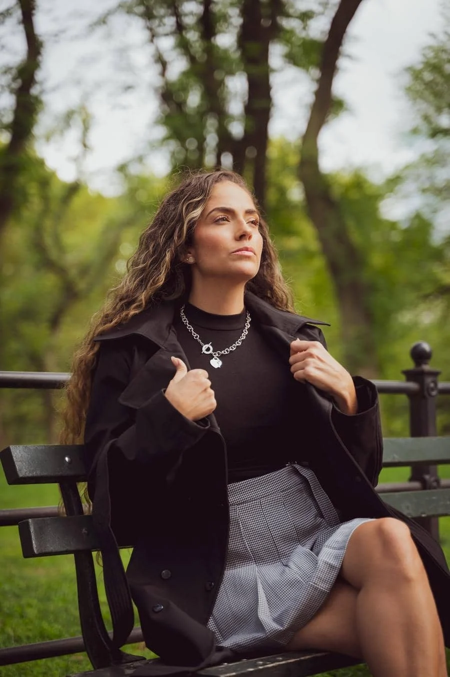 people and lifestyle photography -4 | Pacific Photography Woman sitting on a park bench, wearing a black coat and gray checkered skirt, gazing into the distance with a silver necklace visible.