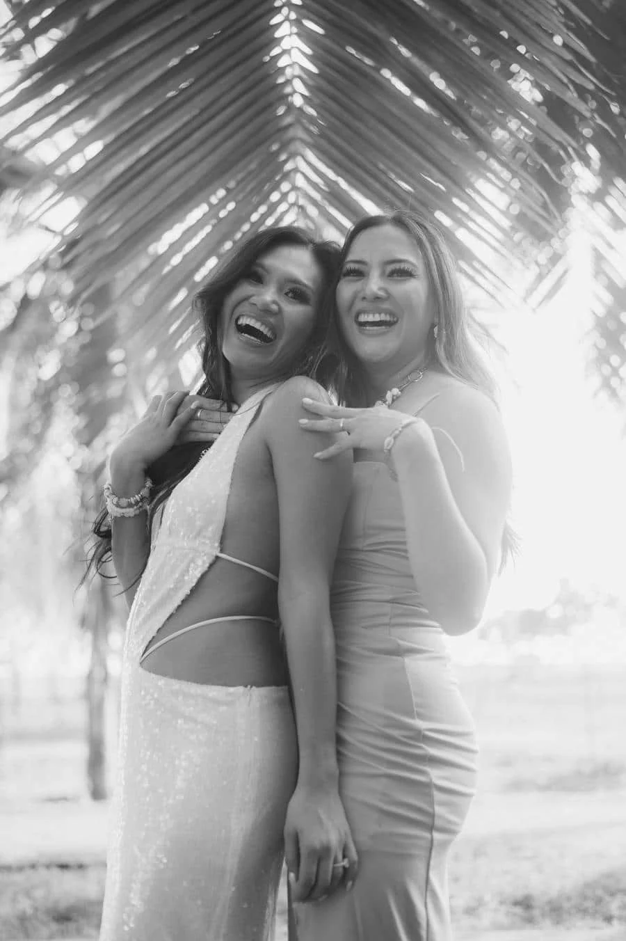 people and lifestyle photography -40 | Pacific Photography Two smiling women in elegant dresses posing together outdoors beneath palm fronds.