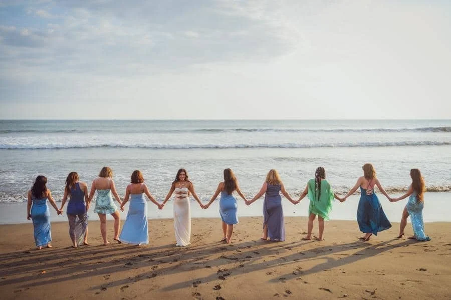 people and lifestyle photography -43 | Pacific Photography Group of women in pastel dresses holding hands on a sandy beach facing the ocean at sunset/sunrise.
