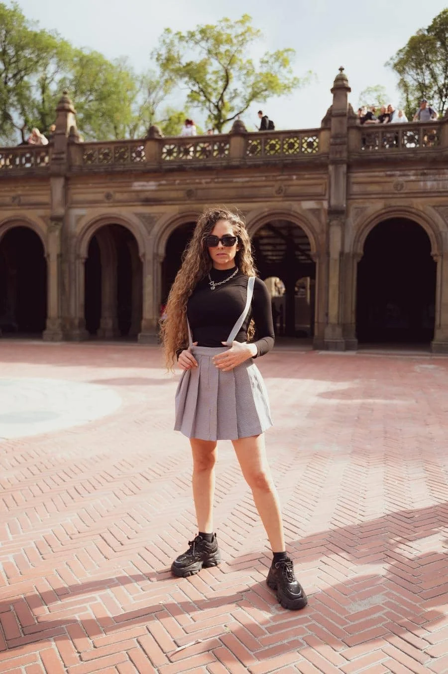 people and lifestyle photography -6 | Pacific Photography Woman posing in black long-sleeve top and gray pleated skirt with suspenders, wearing sunglasses, in a brick-paved plaza with arches behind her.