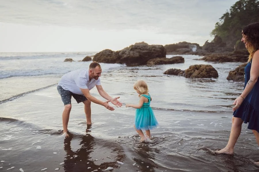 people and lifestyle photography -7 | Pacific Photography Father squats in shallow water to help a toddler in a turquoise dress reach out, as the mother watches from the shore on a rocky beach.