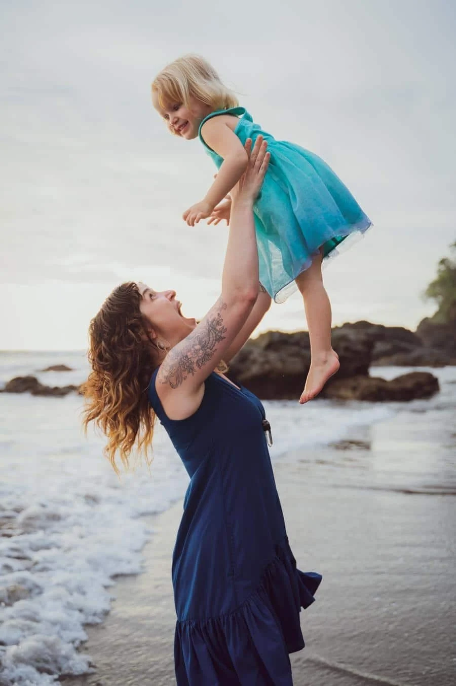 people and lifestyle photography -8 | Pacific Photography Woman in a navy dress lifts a smiling little girl in a turquoise dress above her head on a sandy beach with gentle waves in the background.