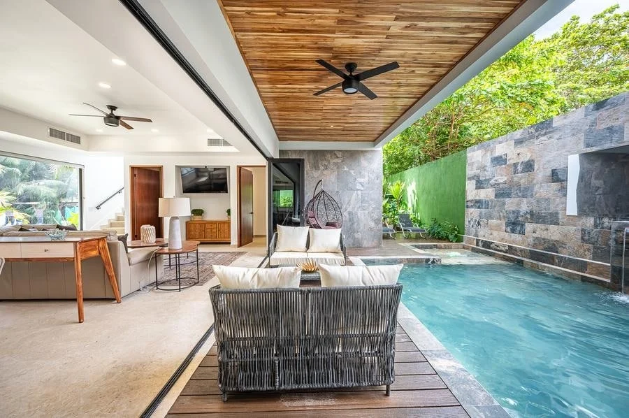 fotografia-para-propiedades-espacios-16 | Pacific Photography Área de estar exterior-interior de planta abierta con piscina, asientos de mimbre y un patio cubierto bajo un techo de madera; un salón, sofá y silla colgante son visibles junto al borde del agua.