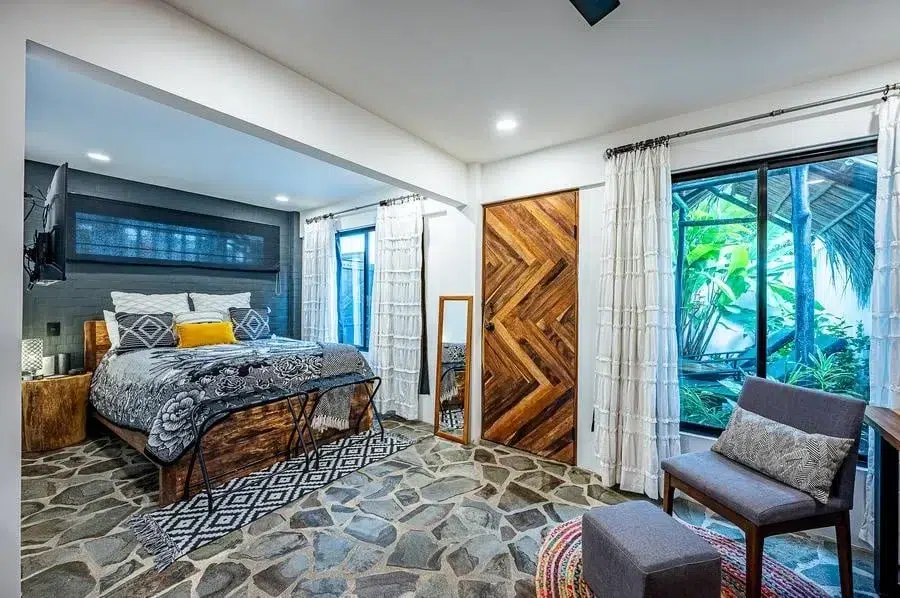 fotografia-para-propiedades-espacios-18 | Pacific Photography Dormitorio moderno con una cama de madera, ropa de cama gris-azul y un suelo de baldosas de piedra; gran ventana con cortinas blancas y plantas tropicales en el exterior.
