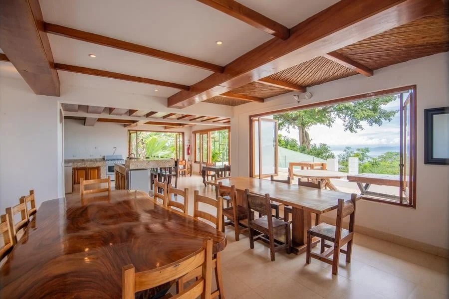 fotografia-para-propiedades-espacios-2 | Pacific Photography Comedor de planta abierta con una larga mesa de madera pulida y varias sillas, vigas de madera expuestas y amplias puertas que enmarcan una ladera verde y una vista lejana al mar de fondo.