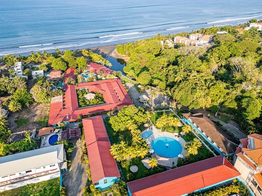 fotografia-para-propiedades-espacios-25 | Pacific Photography Vista aérea de un resort costero con edificios de techos rojos, palmeras y una piscina redonda azul cerca de la playa.