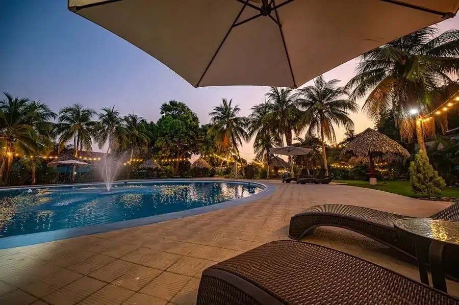 fotografia-para-propiedades-espacios-26 | Pacific Photography Área de piscina estilo resort al anochecer con palmeras, luces de guirnalda y tumbonas de mimbre.