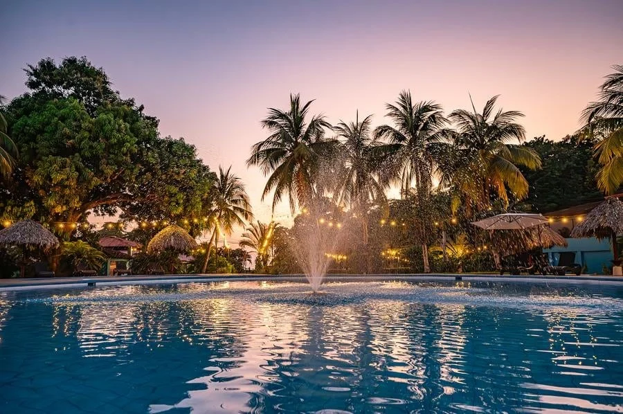 fotografia-para-propiedades-espacios-27 | Pacific Photography Vista al atardecer de una piscina de resort rodeada de palmeras y cabañas con techo de paja, con luces de guirnalda.3d6319